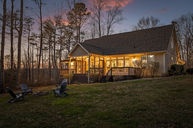 back of house at dusk with a sunroom, roof with shingles, a wooden deck, a fire pit, and a lawn