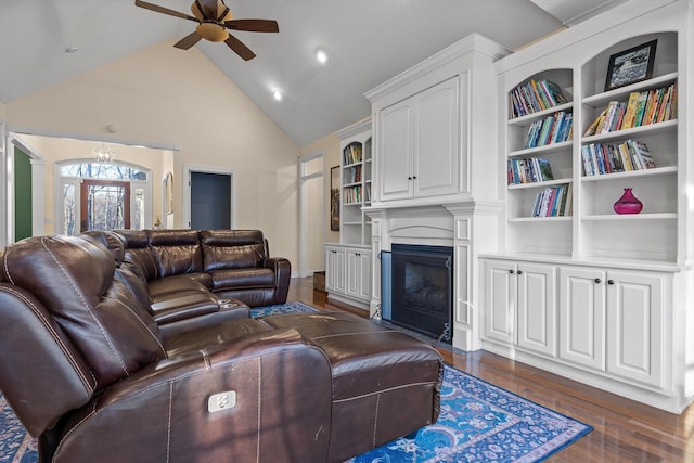 living room featuring dark wood-style flooring, high vaulted ceiling, ceiling fan with notable chandelier, built in shelves, and a glass covered fireplace