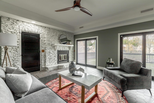 living room with crown molding, baseboards, visible vents, a ceiling fan, and a stone fireplace