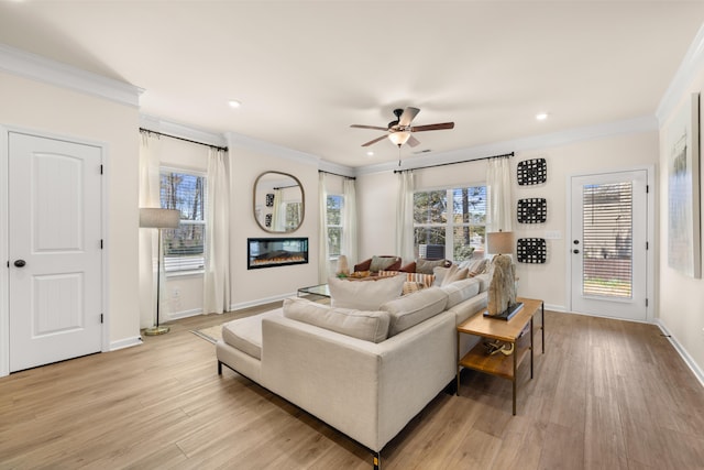 living room featuring light wood-style flooring, baseboards, ornamental molding, a ceiling fan, and a glass covered fireplace