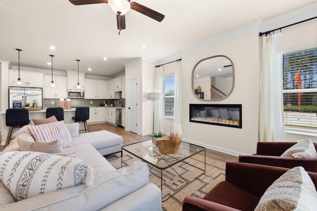 living area featuring crown molding, a ceiling fan, a glass covered fireplace, light wood-type flooring, and baseboards