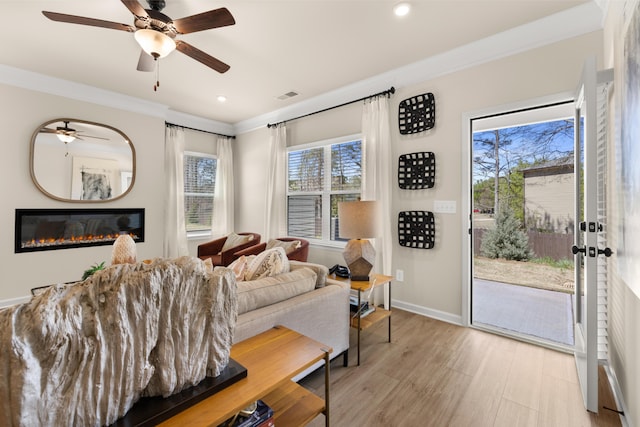 living room featuring baseboards, ornamental molding, a ceiling fan, light wood-type flooring, and a glass covered fireplace