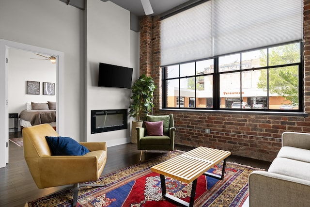living room with a towering ceiling, brick wall, ceiling fan, a glass covered fireplace, and wood finished floors