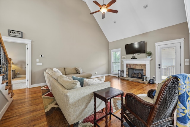 living room featuring high vaulted ceiling, stairway, a fireplace, wood finished floors, and ceiling fan