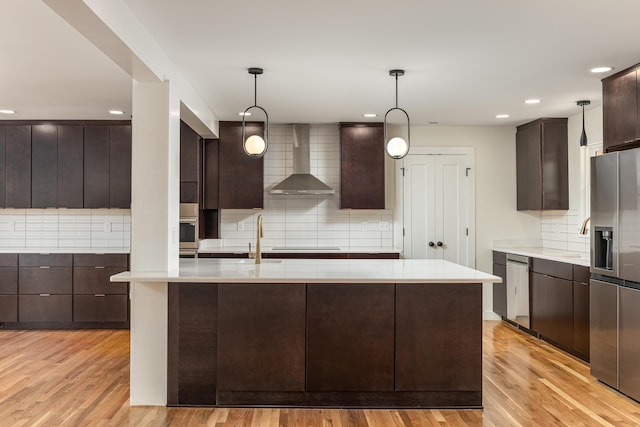 kitchen with dark brown cabinetry, wall chimney range hood, appliances with stainless steel finishes, light countertops, and backsplash