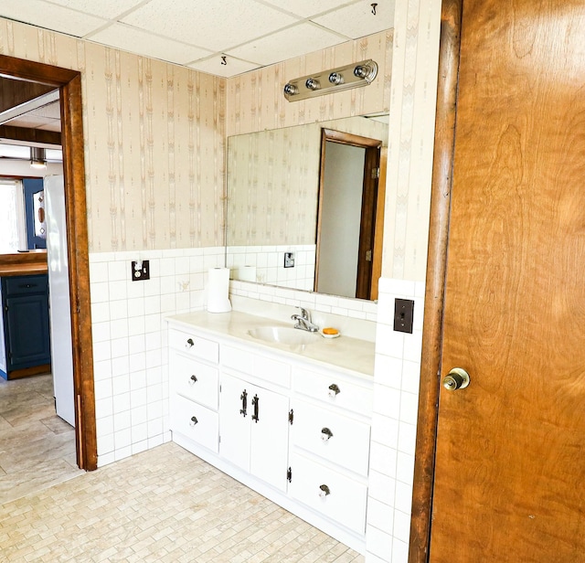 bathroom with a paneled ceiling, vanity, wallpapered walls, wainscoting, and tile walls