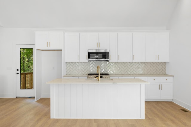 kitchen with stainless steel microwave, backsplash, white cabinets, and an island with sink