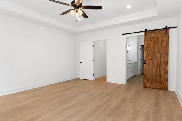 unfurnished bedroom featuring light wood-style flooring, a tray ceiling, a barn door, baseboards, and recessed lighting