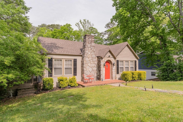 tudor house featuring stone siding, a front lawn, a chimney, and roof with shingles