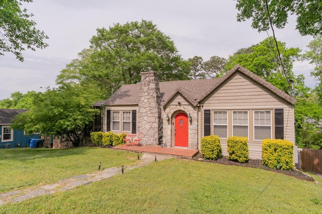 english style home featuring roof with shingles, stone siding, a chimney, fence, and a front yard