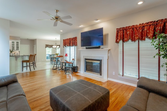 living area with light wood-type flooring, baseboards, ceiling fan with notable chandelier, and a tile fireplace
