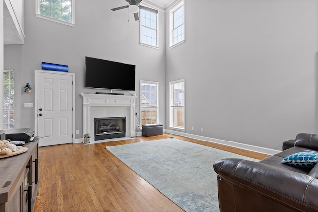 living room featuring a ceiling fan, a tiled fireplace, wood finished floors, baseboards, and a high ceiling