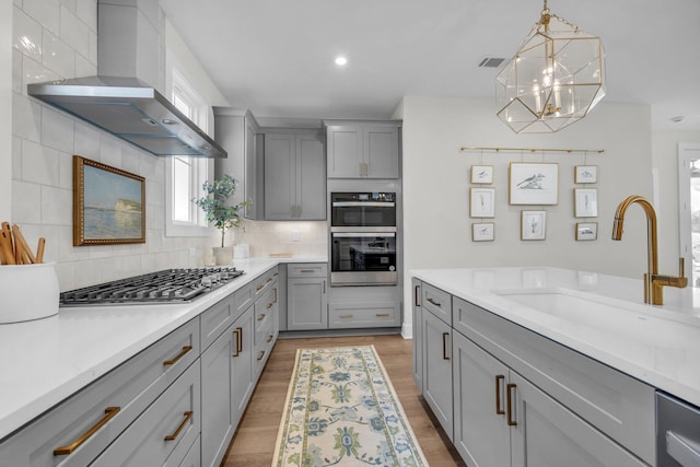 kitchen featuring a sink, stainless steel appliances, gray cabinetry, wall chimney exhaust hood, and backsplash