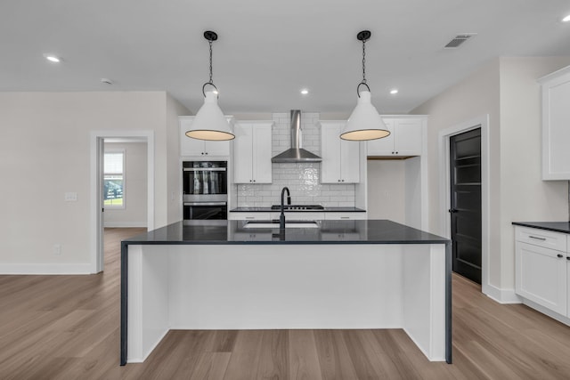 kitchen with double oven, light wood-style flooring, a sink, dark countertops, and wall chimney exhaust hood