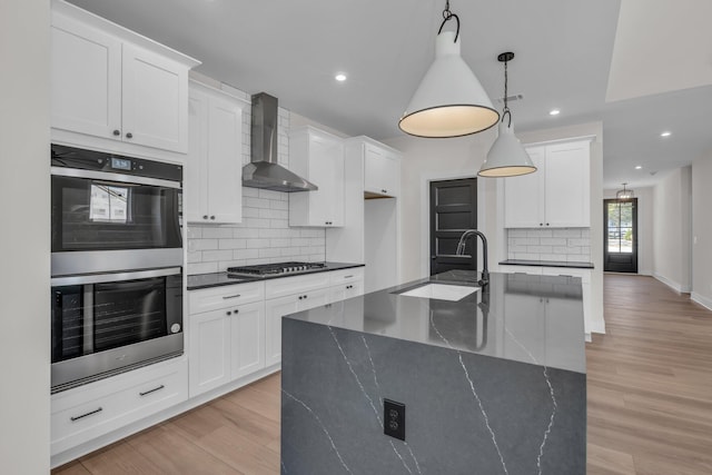 kitchen featuring light wood-style floors, appliances with stainless steel finishes, wall chimney range hood, backsplash, and a sink