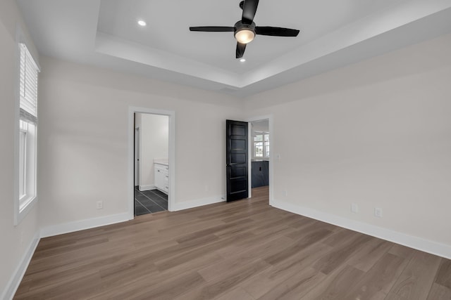 unfurnished bedroom with a raised ceiling, ensuite bathroom, baseboards, recessed lighting, and wood finished floors