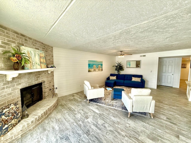 living area with wood finished floors, ceiling fan, a brick fireplace, visible vents, and a textured ceiling