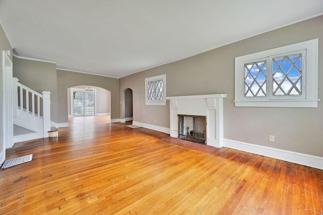 unfurnished living room featuring arched walkways, hardwood / wood-style flooring, a brick fireplace, stairway, and crown molding