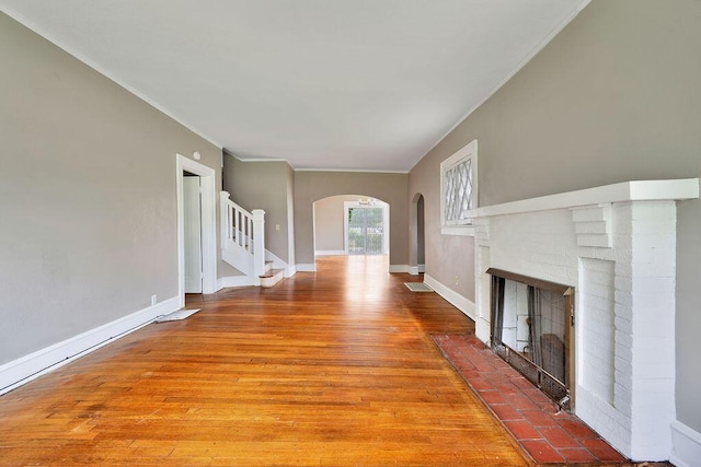 unfurnished living room with a brick fireplace, ornamental molding, arched walkways, stairway, and wood finished floors