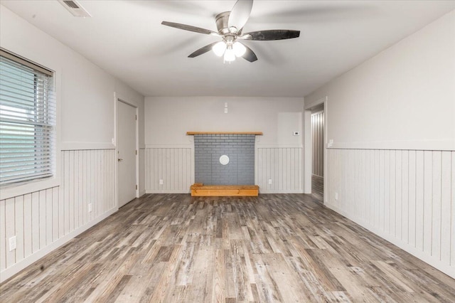 unfurnished living room with visible vents, a brick fireplace, wood finished floors, a wainscoted wall, and a ceiling fan