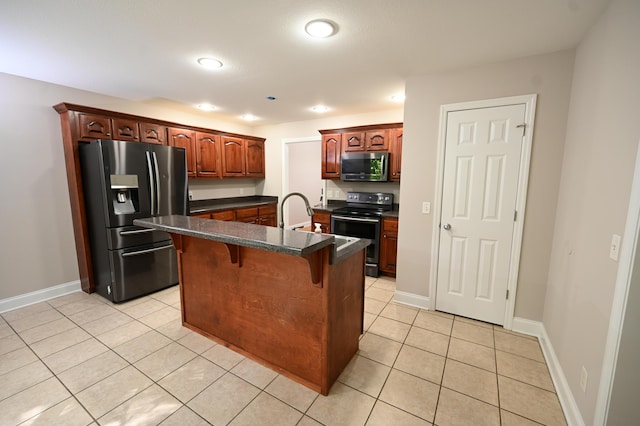 kitchen featuring dark countertops, appliances with stainless steel finishes, light tile patterned floors, a kitchen bar, and a center island with sink