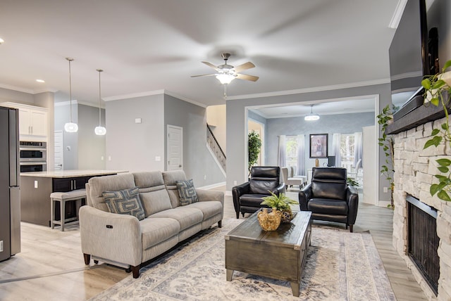 living room featuring crown molding, ceiling fan, light wood finished floors, and a stone fireplace