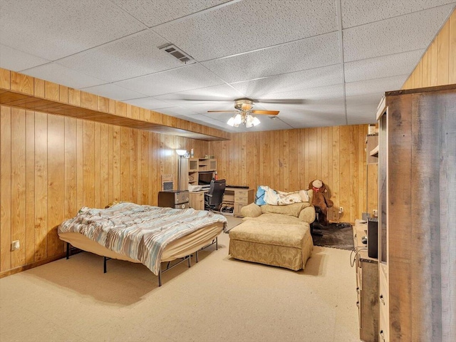 bedroom featuring wooden walls, an office area, carpet floors, a drop ceiling, and ceiling fan