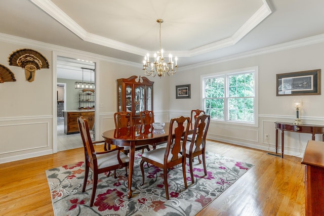 dining space with a chandelier, light wood-style flooring, a raised ceiling, crown molding, and a wainscoted wall