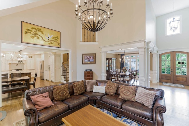 living area featuring a chandelier, a towering ceiling, light wood-style floors, ornate columns, and french doors