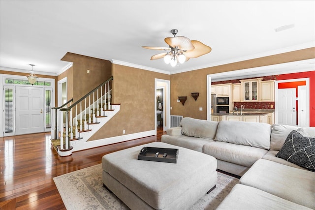 living room featuring dark wood-style floors, baseboards, ornamental molding, a ceiling fan, and stairway