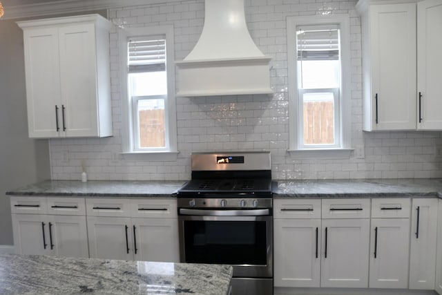 kitchen featuring stainless steel range with gas cooktop, premium range hood, decorative backsplash, white cabinets, and light stone counters