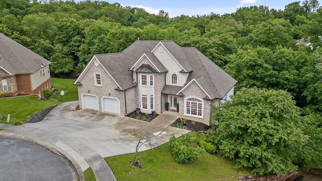 view of front facade with concrete driveway, a view of trees, a front lawn, stone siding, and a shingled roof
