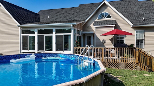 rear view of property with a sunroom, a wooden deck, an outdoor pool, and roof with shingles