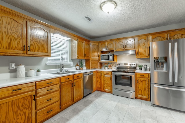 kitchen with stainless steel appliances, a sink, under cabinet range hood, brown cabinetry, and light countertops