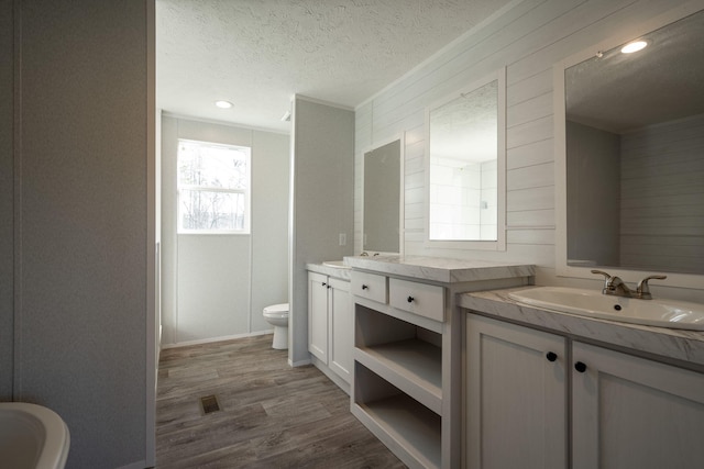 bathroom featuring toilet, wood finished floors, a textured ceiling, two vanities, and wooden walls