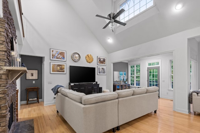living area featuring ceiling fan, high vaulted ceiling, light wood-style flooring, a stone fireplace, and baseboards