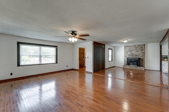 unfurnished living room with ceiling fan, light wood finished floors, a textured ceiling, a brick fireplace, and a wainscoted wall