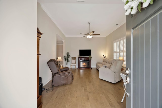 living area featuring ornamental molding, ceiling fan, light wood-type flooring, a glass covered fireplace, and baseboards