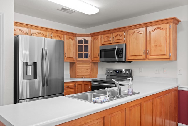 kitchen with stainless steel appliances, light countertops, a sink, brown cabinetry, and glass insert cabinets