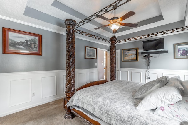 carpeted bedroom featuring a tray ceiling, a wainscoted wall, a textured ceiling, a ceiling fan, and ornamental molding