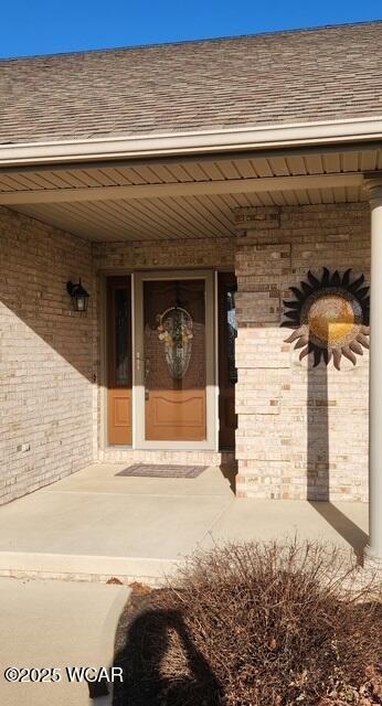 doorway to property with brick siding, a porch, and a shingled roof