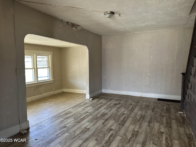 empty room with arched walkways, a textured ceiling, baseboards, and wood finished floors