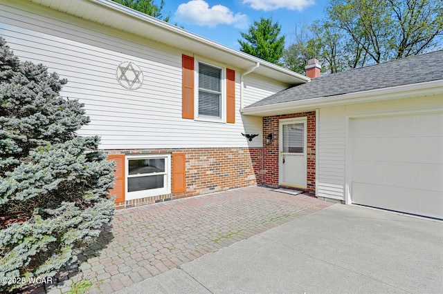 entrance to property featuring driveway, a chimney, roof with shingles, brick siding, and an attached garage