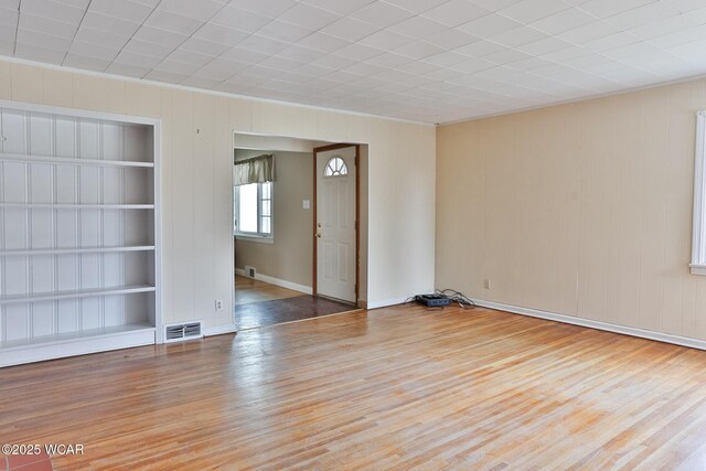 empty room featuring light wood-type flooring, built in shelves, visible vents, and ornamental molding