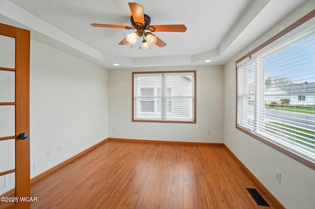 empty room with a tray ceiling, visible vents, light wood-type flooring, baseboards, and ceiling fan