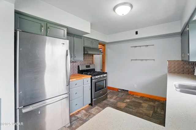 kitchen featuring under cabinet range hood, stone finish floor, appliances with stainless steel finishes, backsplash, and baseboards