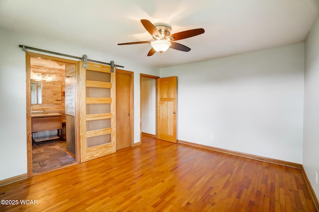 unfurnished bedroom with a barn door, ceiling fan, baseboards, light wood finished floors, and ensuite bath