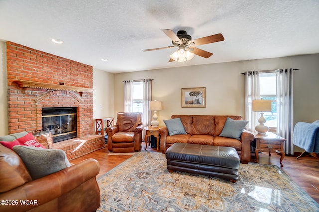 living room featuring a brick fireplace, wood-type flooring, a textured ceiling, and ceiling fan