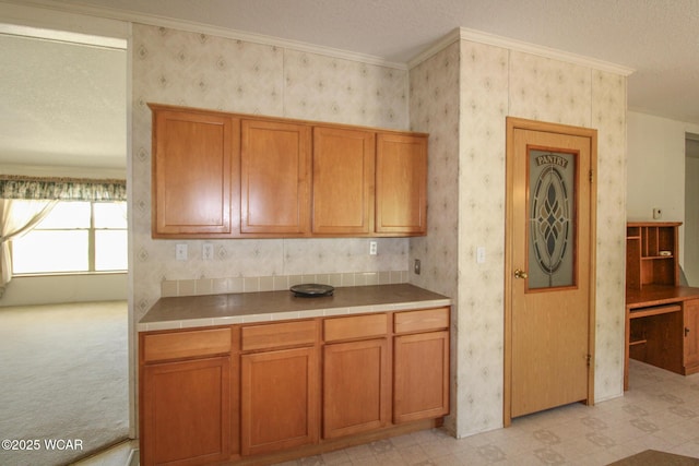 kitchen with wallpapered walls, crown molding, tile countertops, and brown cabinetry
