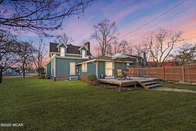 back of house at dusk featuring fence, a wooden deck, central AC, a chimney, and a lawn